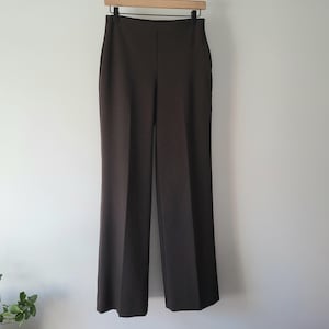 May include: Dark brown, high-waisted trousers hanging on a wooden hanger. The pants have a wide leg and a simple, classic design. The trousers are made of a smooth, solid-colored fabric.