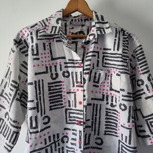 Puede incluir: Camisa blanca con un patr&oacute;n geom&eacute;trico negro y detalles rosas. La camisa tiene mangas cortas, un bolsillo en el pecho y botones rosas. Cuelga de una percha de madera.