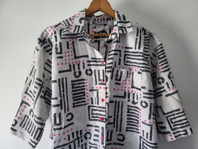 Puede incluir: Camisa blanca con un patr&oacute;n geom&eacute;trico negro y detalles rosas. La camisa tiene mangas cortas, un bolsillo en el pecho y botones rosas. Cuelga de una percha de madera.