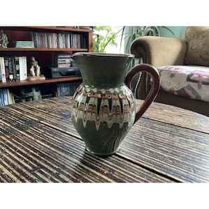 May include: A green ceramic pitcher with a brown handle and a decorative pattern of wavy lines in white, brown, and orange. The pitcher is on a wooden table.