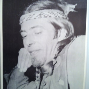 May include: Black and white photo of John Mayall, a musician, with a headband and long hair. He is looking down and has his hand on his chin.