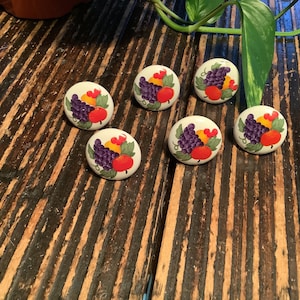 May include: Six round cabinet knobs with a hand-painted fruit design. Each knob features a cluster of purple grapes, a red apple, yellow fruit, and green leaves on a cream background. The knobs are arranged on a wooden surface.