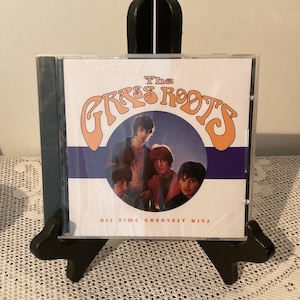 Puede incluir: Una caja de CD con la banda "The Grass Roots" y el texto "All Time Greatest Hits". La portada del álbum tiene un diseño retro con los miembros de la banda en una foto circular. El nombre de la banda está en letras naranjas y blancas.