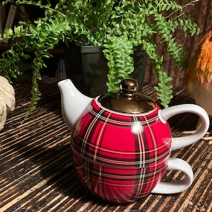 May include: A red and black tartan teapot with a gold lid and white handle. The teapot is sitting on a wooden table with two decorative chickens on either side.