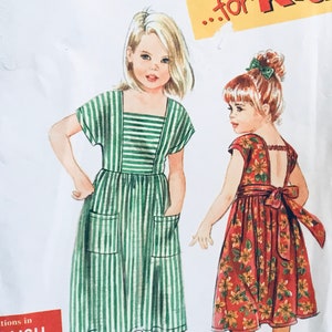May include: A vintage sewing pattern for two children's dresses. The pattern features a green and white striped dress with pockets and a red floral dress with a bow at the back. The pattern is titled "...for Kids".