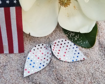 May include: A pair of silver leaf-shaped earrings with a pattern of red, white, and blue stars. The earrings are displayed on a sandy surface, with a small American flag and white flower in the background.