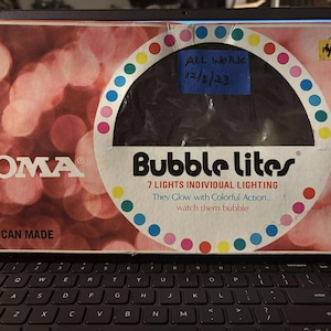 Può includere: Una scatola vintage di luci di Natale Noma Bubble Lites. La scatola è rossa e bianca con un'immagine delle luci al centro. Il testo sulla scatola recita "Bubble Lites" e "7 Lights Individual Lighting". La scatola dice anche "All American Made" e "Pre-Tested".