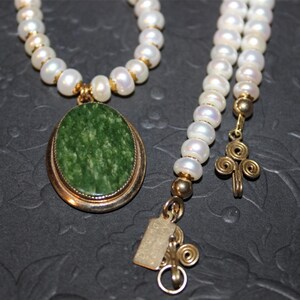 May include: A white pearl necklace with a green stone pendant. The pendant is set in gold and has a detailed design. The necklace is finished with a gold clasp and a small gold charm.