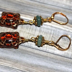 May include: A pair of gold earrings with a brown turtle charm and a green glass bead. The earrings have a hook closure.