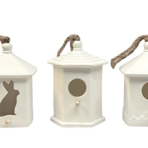 May include: Three white ceramic birdhouses with a rustic twine hanger. One birdhouse has a bunny silhouette cutout, another has a round opening, and the third has a scalloped opening.