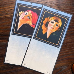 May include: Two vintage art deco prints featuring portraits of women with vibrant makeup and headscarves. The portraits are framed in black with gold accents, set against a light blue background. The prints have a small hole at the top for hanging.