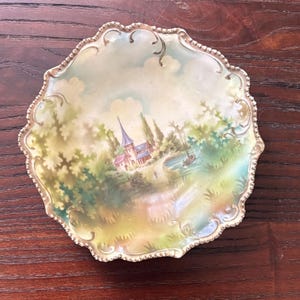 May include: A decorative, scalloped-edge plate with a painted landscape scene. The artwork depicts a church with a tall steeple, trees, and a small boat on a lake. The plate has a silver-colored trim.