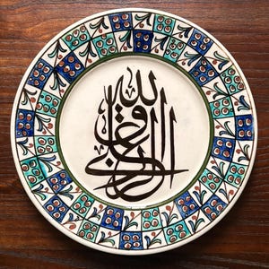 May include: A decorative ceramic plate with a white center featuring black Arabic calligraphy. The rim is adorned with a repeating pattern of blue, green, and brown squares and floral motifs, set against a white background.