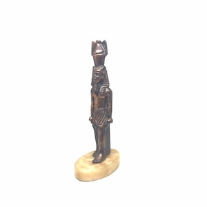 May include: A small bronze statue of an Egyptian pharaoh standing on a beige oval base. The pharaoh is wearing a traditional headdress and kilt.