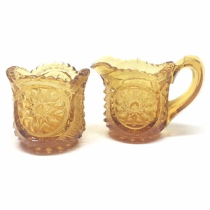 May include: A set of two amber glass creamer and sugar bowls with a decorative, starburst pattern. The bowls have a scalloped edge and a textured base.