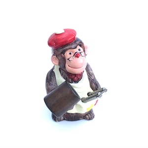 Könnte beinhalten: Eine Keramikfigur eines Affen, der einen roten Barett trägt und einen Pinsel und einen Holzhammer hält. Der Affe trägt eine weiße Schürze mit einem gelben und roten Farbklecksmuster.