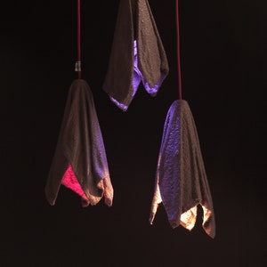 Pu&ograve; includere: Tre lampadari sospesi in tessuto con una superficie strutturata. I lampadari hanno la forma di un fiore e sono illuminati da luci di diversi colori. Le luci sono viola, rosa e gialle.