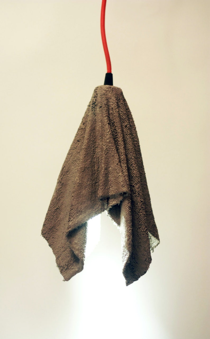 Pu&ograve; includere: Una lampada a sospensione in cemento grigio con un cavo rosso che pende dal soffitto.