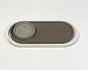 May include: An oval-shaped tray featuring a white outer border and a dark gray inner surface. A smaller, circular gray coaster is positioned on the left side of the tray, creating a layered, multi-toned design with smooth, solid edges.