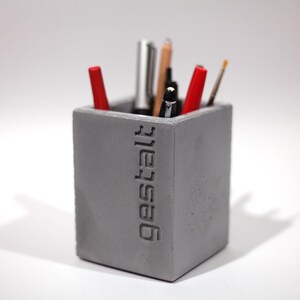 May include: A gray concrete pencil holder with the word "Gestalt" embossed on the side. The holder is filled with various pens and pencils, including red, black, and silver.