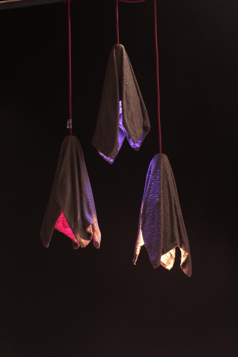 Pu&ograve; includere: Tre lampadari sospesi in tessuto con una superficie strutturata. I lampadari hanno la forma di un fiore e sono illuminati da luci di diversi colori. Le luci sono viola, rosa e gialle.