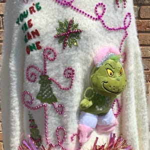 May include: Cream-coloured, fluffy Christmas jumper with pink and green decorations. Includes the text "You're the one" and a Grinch plush toy. Adorned with pink beads, tinsel, and floral details. Festive and holiday-themed.