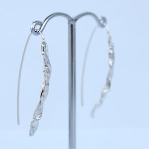 Puede incluir: Un par de pendientes plateados con un diseño texturizado único. Cada pendiente presenta un alambre curvado que se conecta a un colgante retorcido de forma orgánica. Los pendientes se exhiben en un soporte plateado sobre un fondo azul claro.