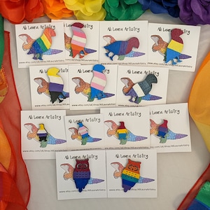 May include: A collection of 10 colourful wooden pins featuring different pride flags. The pins are shaped like lizards and owls. The pins are displayed on white cards with the text "Ali Laura Artistry" and a website address.
