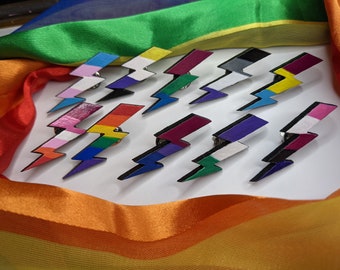May include: A set of 12 lightning bolt pins, each painted with a different pride flag design. The pins are arranged in a semi-circle on a white surface.