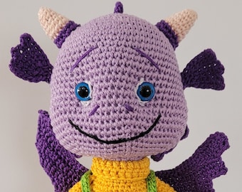 Peut inclure: Un dragon violet en crochet portant une chemise jaune et une salopette verte avec des boutons en forme de cœur verts. Le dragon a un visage souriant avec des yeux bleus.