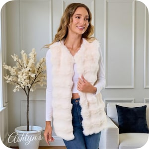 May include: Cream-colored faux fur vest. The vest has a vertical, ribbed design and a V-neck. The model is wearing a white long-sleeve shirt and blue jeans. The vest is a stylish outerwear piece.