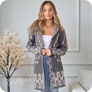 May include: A long, open-front gray cardigan with a patterned design. The cardigan features a hood and a geometric pattern in shades of white, orange, and blue. The text "Ashlyn's" is visible in the lower left corner.