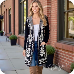 May include: A long black cardigan with a geometric pattern in white and tan. The cardigan has long sleeves, pockets, and a striped collar. The model is wearing blue jeans and brown boots. The cardigan is open, revealing a white top.