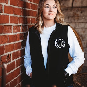 May include: A woman wearing a black fleece vest with a white long-sleeved shirt underneath. The vest has a white monogram on the front that reads "ROL".