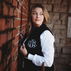 May include: A woman wearing a black fleece vest with a white monogram on the front, a white long-sleeved shirt, and dark hair. She is standing next to a brick wall.