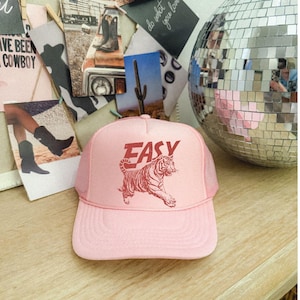 Puede incluir: Gorra de camionero rosa claro con la palabra "EASY" en rojo sobre un gr&aacute;fico de tigre rojo. La gorra tiene una parte trasera de malla y una visera curva. La gorra est&aacute; sobre una superficie de madera.
