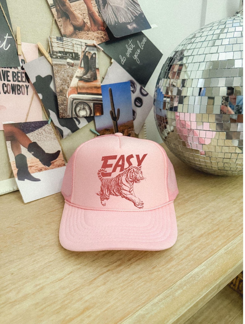 Puede incluir: Gorra de camionero rosa claro con la palabra "EASY" en rojo sobre un gr&aacute;fico de tigre rojo. La gorra tiene una parte trasera de malla y una visera curva. La gorra est&aacute; sobre una superficie de madera.