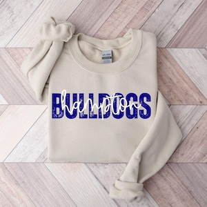 May include: A beige sweatshirt with the word "BULLDOGS" in blue block letters and "hampton" in white script. The sweatshirt has a crew neck and long sleeves, with the left sleeve folded.