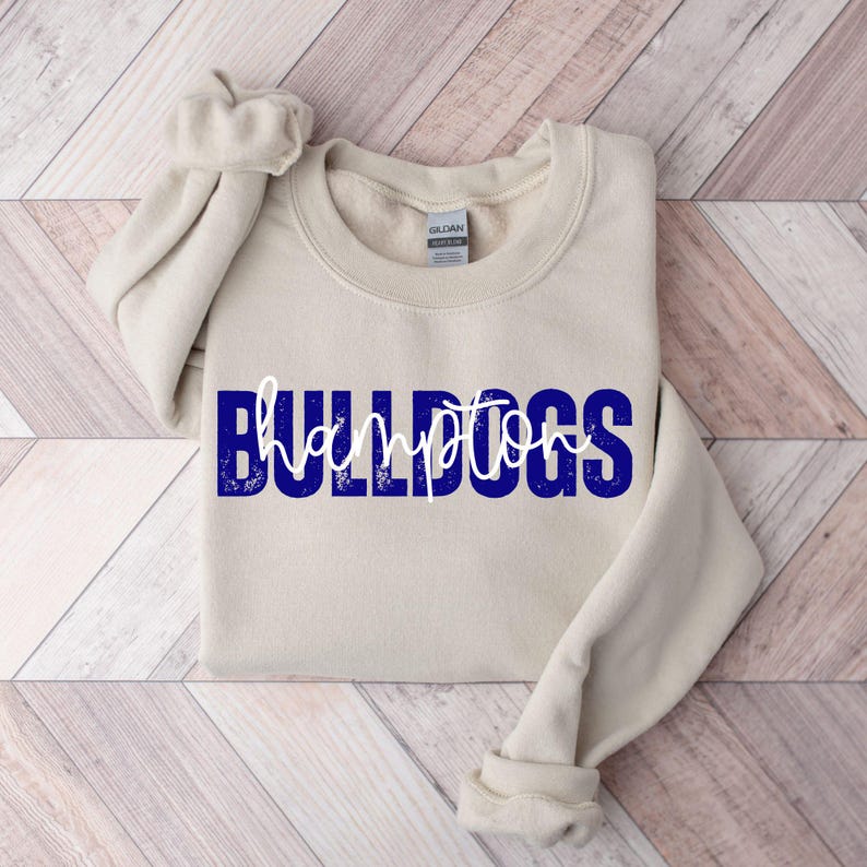 May include: A beige sweatshirt with the word "BULLDOGS" in blue block letters and "hampton" in white script. The sweatshirt has a crew neck and long sleeves, with the left sleeve folded.