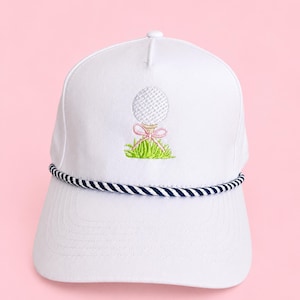 May include: White golf hat with a navy and white rope brim. The hat features an embroidered golf ball design with a pink bow and green grass. A stylish accessory for golf enthusiasts.