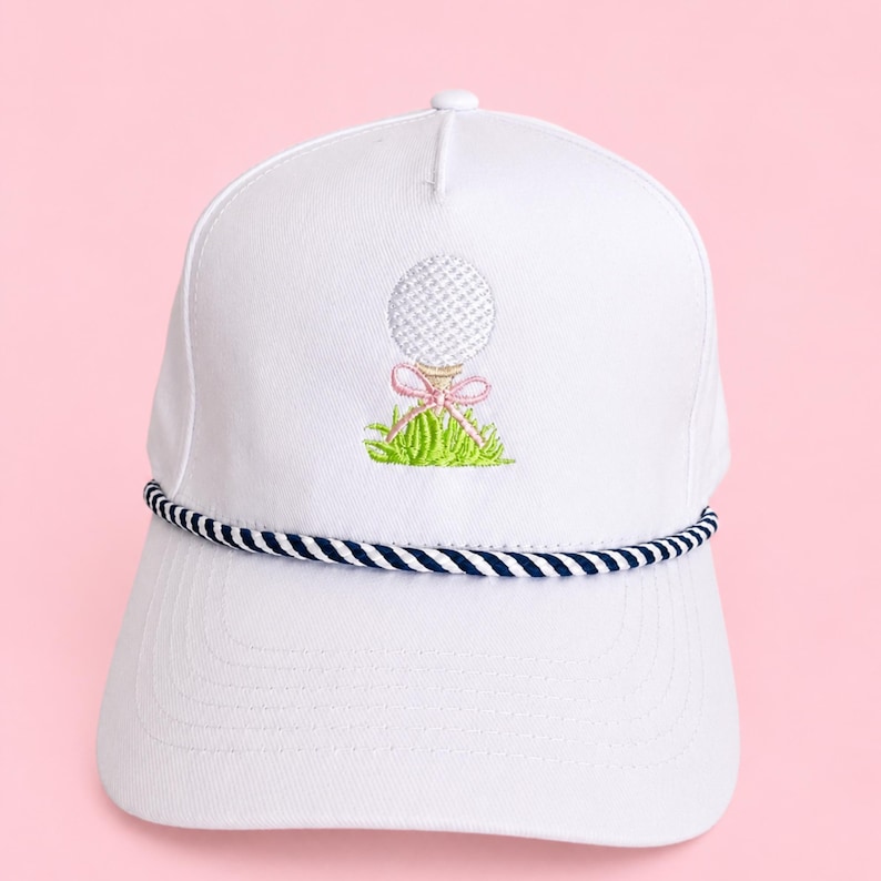 May include: White golf hat with a navy and white rope brim. The hat features an embroidered golf ball design with a pink bow and green grass. A stylish accessory for golf enthusiasts.