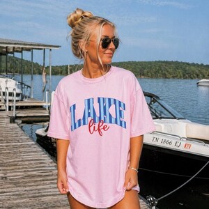 May include: A light pink t-shirt with the words "LAKE life" in blue and red lettering. The shirt is worn by a person on a wooden dock near a lake, with a boat in the background.