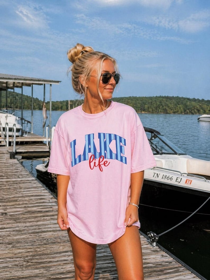 May include: A light pink t-shirt with the words "LAKE life" in blue and red lettering. The shirt is worn by a person on a wooden dock near a lake, with a boat in the background.