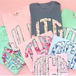 May include: A collection of pastel-colored t-shirts, including pink, blue, and green, each featuring a floral or animal print monogram design. The shirts are neatly folded and arranged on a pink surface.