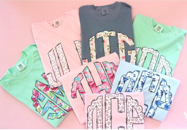 May include: A collection of pastel-colored t-shirts, including pink, blue, and green, each featuring a floral or animal print monogram design. The shirts are neatly folded and arranged on a pink surface.