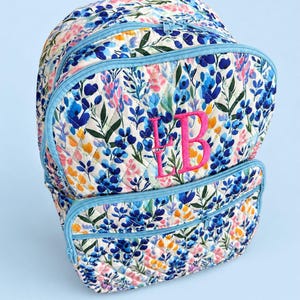 May include: A quilted backpack featuring a white base with a colorful floral pattern in shades of blue, pink, and yellow. The bag has light blue zippers and trim, a top handle, and the embroidered pink monogram letters LB on the front pocket.