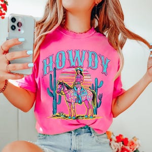 May include: A bright pink t-shirt with the word "HOWDY" in turquoise lettering. The shirt features a graphic of a cowgirl riding a horse, with cacti and a sunset in the background. The design is colorful and eye-catching.
