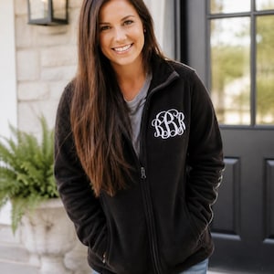 May include: Black fleece zip-up jacket with a white monogram on the left chest. The jacket has a high collar, two front pockets, and a full-length zipper. The person in the image is wearing a gray t-shirt and blue jeans.