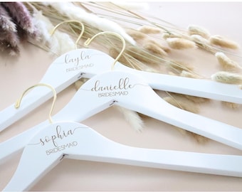 May include: Three white wooden hangers with gold hooks. Each hanger has a different name printed on it in gold script: "layla bridesmaid", "danielle bridesmaid", and "sophia bridesmaid".