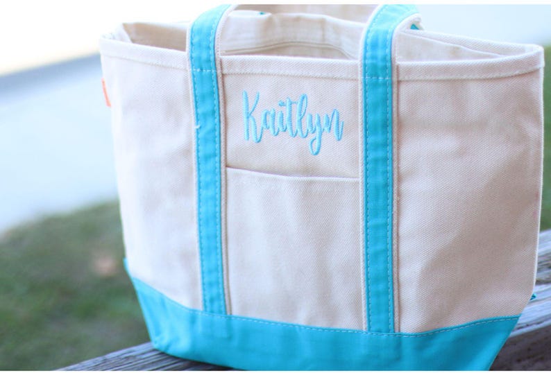 May include: A white canvas tote bag with blue trim and a personalized embroidered name, "Kaitlynn".